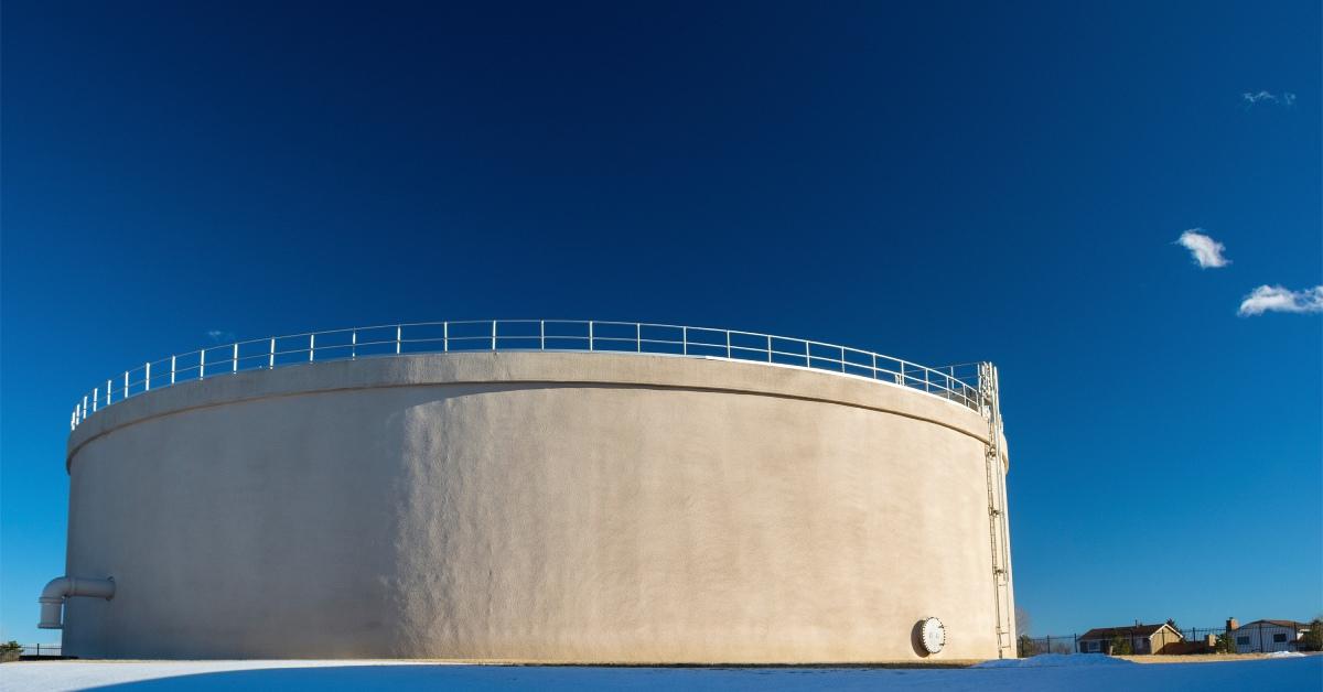 A large water tank sits on a patch of land covered in snow. The tank is dark gray and the sky behind it is blue.