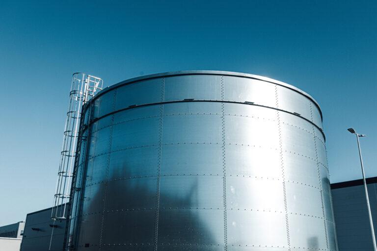 A metal water tank reflects the sun off of its surface. There's a building and light pole in the background.