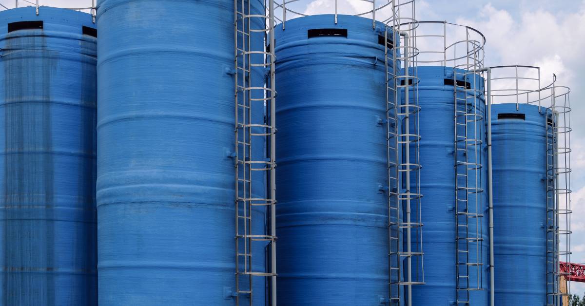 Five tall blue chemical storage containers are sitting next to each other with silver ladders attached to the sides.